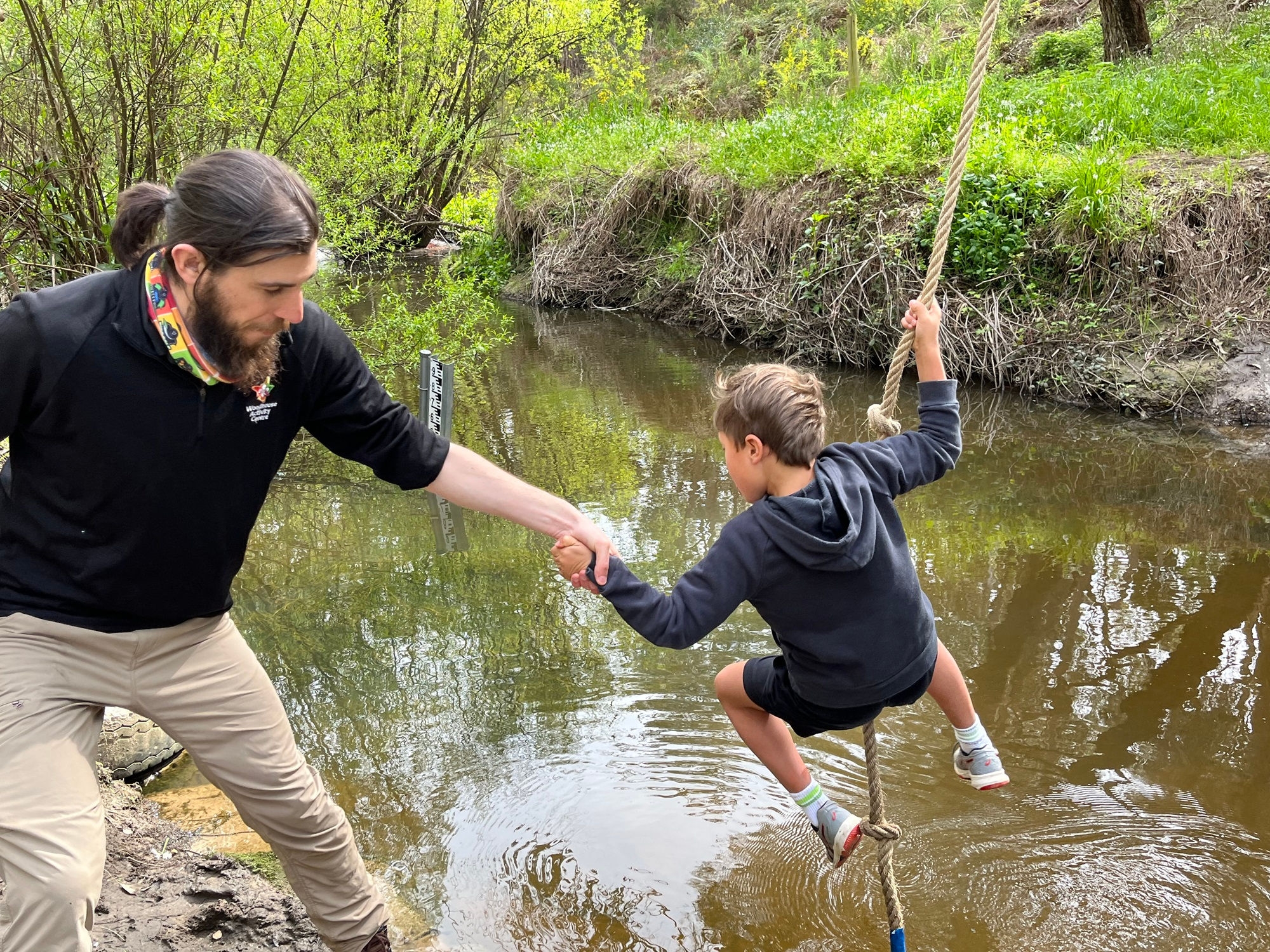 A man on the left helping a young boy holding on to a rope to swing over a creek.