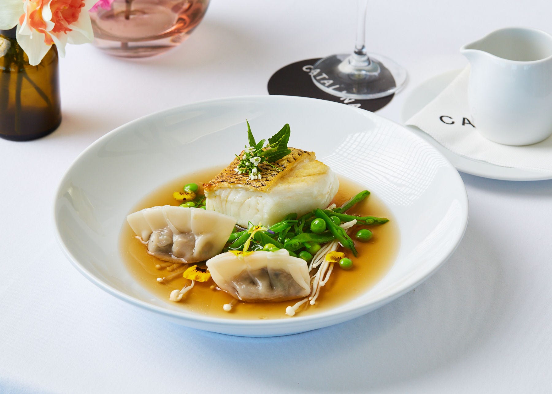A plate with a fillet of toothfish, two dumplings, shelled and unshelled peas, enoki mushrooms and edible flowers, in a light broth.