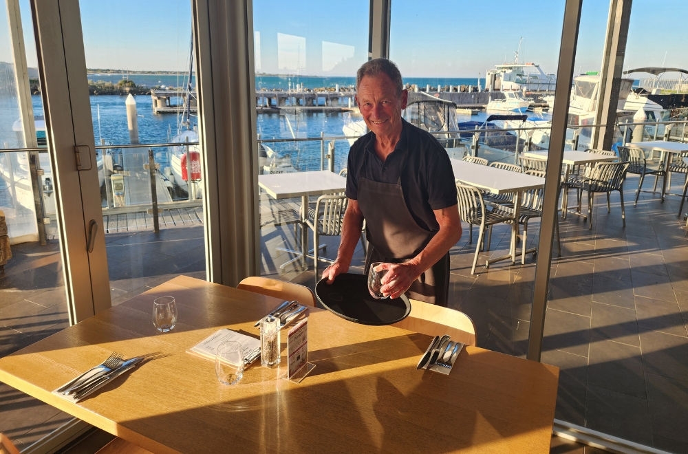 A man standing in front of a table holding a tray with a glass. Behind him is a floor to ceiling window with a water view.